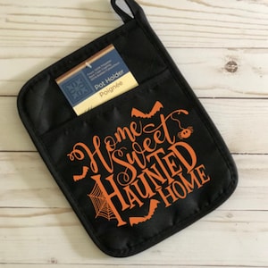 May include: Black pot holder with the text "Home Sweet Haunted Home" in orange script. The design includes bats and a spider web. A tag in the pocket reads "Pot Holder" and "Poignée".