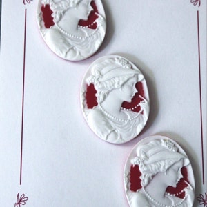 May include: Three white cameo buttons with a red background. Each button features a woman's profile wearing a hat and a pearl necklace. The buttons are labeled "Donna May's Lovely Buttons".
