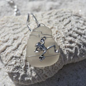 Peut inclure: Un pendentif en forme de grenouille argentée suspendu à une chaîne argentée. Le pendentif est en verre transparent avec un motif de fil argenté. Le pendentif repose sur une surface blanche de type corail.