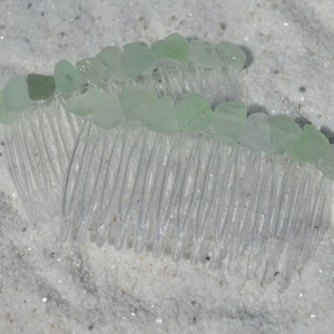 May include: A clear plastic hair comb with light green sea glass pieces attached to the teeth. The comb is lying on a white sandy surface.