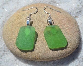 May include: A pair of green sea glass earrings with silver hooks. The earrings are displayed on a light brown rock.