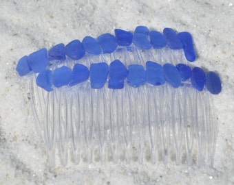 May include: A pair of clear plastic hair combs decorated with light blue sea glass. The combs are lying on a white sandy surface.