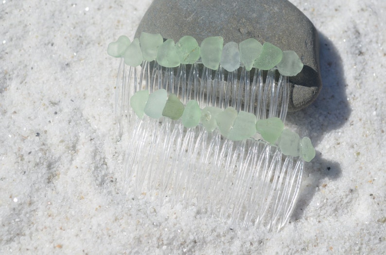 May include: Two clear plastic hair combs decorated with light green sea glass. The combs are lying on a white sandy surface.