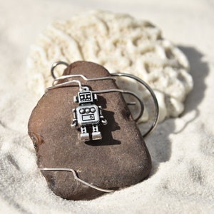 May include: A silver robot charm is attached to a brown rock wrapped in silver wire. The rock is sitting on a white sandy surface.