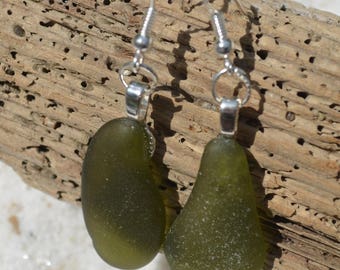 May include: A pair of olive green sea glass earrings with silver-colored hooks. The teardrop-shaped glass pieces have a frosted texture and are suspended from simple, elegant ear wires. The earrings rest on a piece of driftwood.