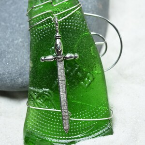 May include: A handcrafted pendant featuring a piece of green glass with a silver sword charm. The glass is wrapped with silver wire. The pendant is displayed on a white surface with a gray stone in the background.