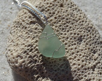 May include: A light green sea glass pendant wrapped in silver wire, hanging from a silver chain. The pendant is resting on a piece of coral.