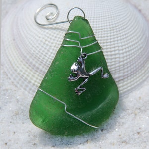 Peut inclure: Ornement artisanal en verre de mer vert, enveloppé de fil d'argent. Un petit charm en forme de grenouille argentée est attaché. Il est doté d'une boucle pour être suspendu. Fond : coquillage et sable.