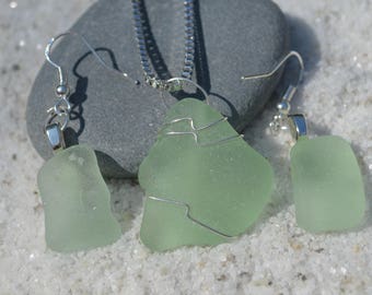 May include: A sea glass jewelry set featuring a necklace and earrings. The necklace has a large, light green sea glass pendant wrapped in silver wire, suspended from a silver chain. The earrings have matching sea glass pieces. The set is displayed on a gray stone.