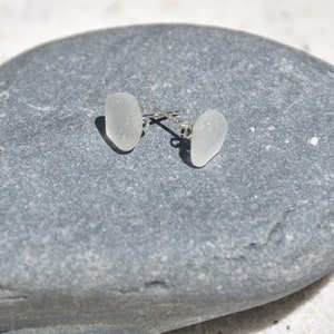 Pode incluir: Um par de brincos de vidro do mar branco com postes de prata. Os brincos estão sobre um fundo de pedra cinza.