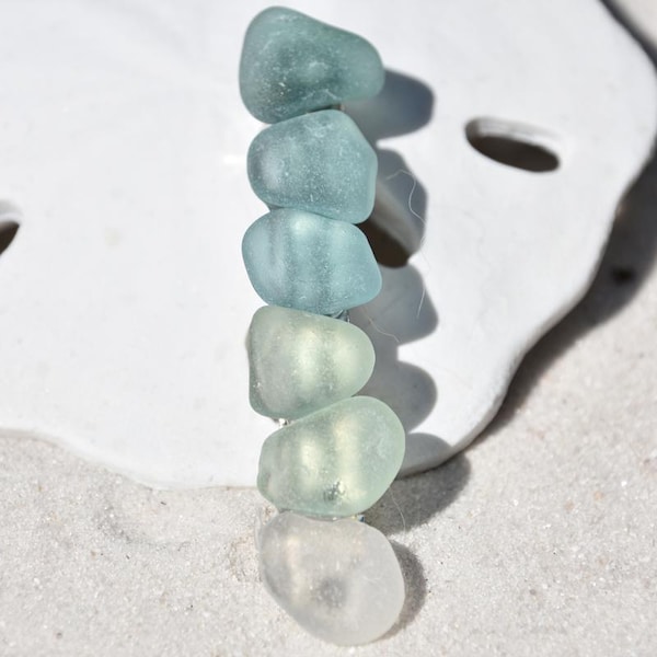May include: A collection of sea glass beads in shades of blue and green, arranged in a gradient from dark to light.
