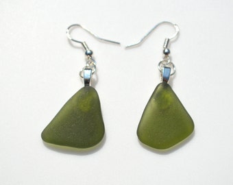May include: A pair of olive green sea glass earrings. Each earring features a triangular piece of sea glass suspended from a silver-colored fish hook ear wire. The sea glass has a frosted appearance and a slightly irregular shape.