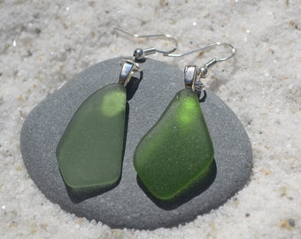 May include: A pair of handcrafted earrings featuring sea glass pendants in shades of green. The sea glass is mounted on silver-colored hooks, creating a unique and natural jewelry piece. The earrings are displayed on a gray stone against a backdrop of white sand.