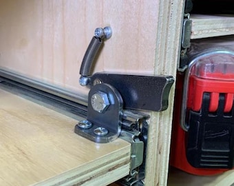 May include: Close-up of a metal drawer slide with a spring-loaded latch attached to a wooden cabinet. The latch is in the open position, showing the metal spring and the metal arm that holds the latch in place.