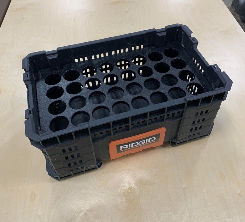 K&ouml;nnte beinhalten: Schwarze Plastik-Aufbewahrungsbox mit einem roten und schwarzen RIDGID-Etikett. Die Box hat ein Gittermuster und einen herausnehmbaren Einsatz mit 24 runden L&ouml;chern.