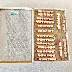 May include: Open cardboard box containing multiple sets of artificial teeth. The teeth are arranged on red and clear plastic strips. The left side of the box contains bubble wrap and a small blue label that reads "FINAL INSPECTION".