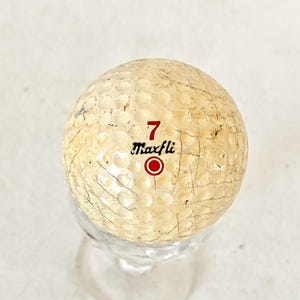 May include: A vintage golf ball with the number 7 and the brand name "Maxfli" printed in red. The ball has a dimpled surface and a worn, off-white color, suggesting it has been used.
