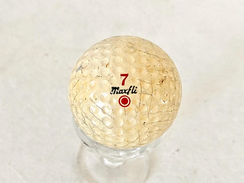 May include: A vintage golf ball with the number 7 and the brand name "Maxfli" printed in red. The ball has a dimpled surface and a worn, off-white color, suggesting it has been used.