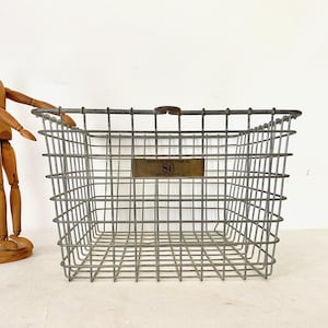 May include: A rectangular metal wire basket with a rectangular brass label that reads "SPECIAL WIRE FORM CO. 86". The basket is gray and has a wooden handle. A wooden artist's mannequin stands to the left of the basket.