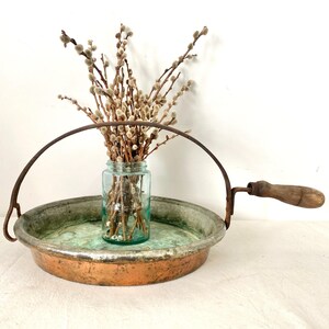 May include: An antique copper and metal tray with a handle and a wooden grip. A clear glass jar filled with pussy willow branches sits in the center of the tray. The tray has a green and copper patina.