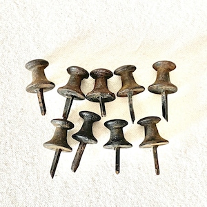 May include: A collection of nine metal push pins featuring a weathered, dark brown and oxidized finish. Each pin has a rounded, mushroom-shaped head and a sharp, pointed metal spike extending from the center, showing signs of age and patina.