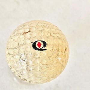 May include: A used golf ball with a cracked, textured surface. The ball is off-white with a black and red logo. The dimpled surface shows signs of wear and age, indicating previous use on a golf course.