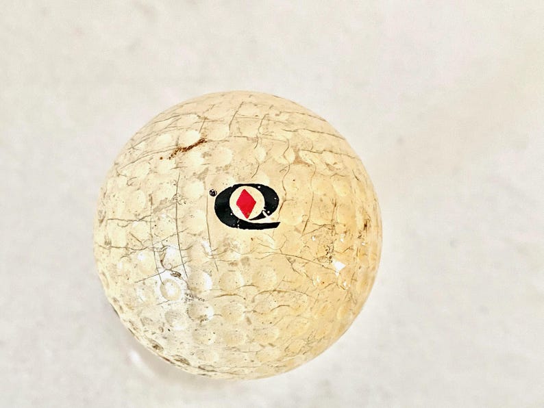 May include: A used golf ball with a cracked, textured surface. The ball is off-white with a black and red logo. The dimpled surface shows signs of wear and age, indicating previous use on a golf course.