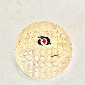 May include: A used golf ball with a worn, textured surface. The ball is off-white with a black and red logo. The ball has visible scuff marks and small brown spots, indicating prior use. The dimpled surface is typical of golf balls.
