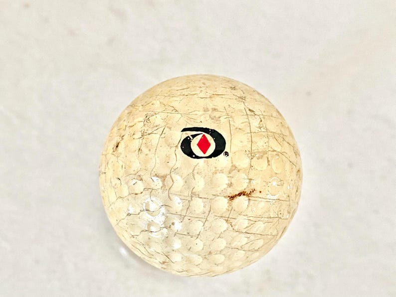 May include: A used golf ball with a worn, textured surface. The ball is off-white with a black and red logo. The ball has visible scuff marks and small brown spots, indicating prior use. The dimpled surface is typical of golf balls.