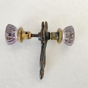 May include: Antique door handle with two lavender glass knobs and brass hardware. The handle has a decorative, ornate design and a weathered, vintage appearance. The glass knobs have a ribbed texture.
