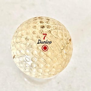May include: A used Dunlop golf ball, featuring the number 7 in red, the brand name, and a red circle. The ball has a dimpled surface and visible wear and tear.