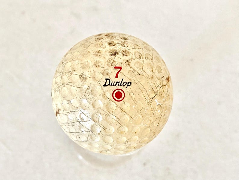 May include: A used Dunlop golf ball, featuring the number 7 in red, the brand name, and a red circle. The ball has a dimpled surface and visible wear and tear.