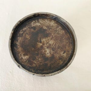 May include: A circular ceramic dish with a dark, mottled interior and a grey rim. The interior has a textured, aged appearance with shades of brown and grey. The rim is a lighter grey, creating a contrast with the darker centre.