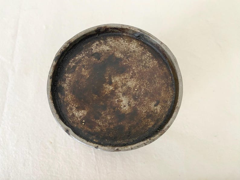 May include: A circular ceramic dish with a dark, mottled interior and a grey rim. The interior has a textured, aged appearance with shades of brown and grey. The rim is a lighter grey, creating a contrast with the darker centre.