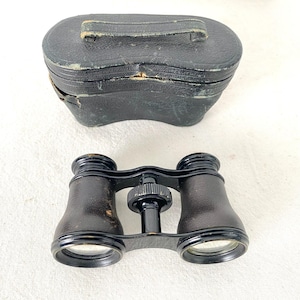 May include: Antique black opera glasses with a matching black leather case. The binoculars have a central focus wheel and are made of metal and leather. The case has a handle and shows signs of wear.