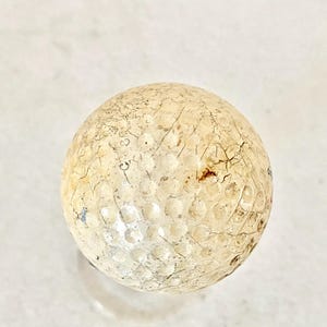 May include: A vintage golf ball with a weathered appearance. The ball is off-white with visible cracks and imperfections across its surface. The dimpled design is still apparent, showing signs of age and use. A classic sports collectible.