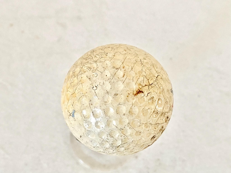 May include: A vintage golf ball with a weathered appearance. The ball is off-white with visible cracks and imperfections across its surface. The dimpled design is still apparent, showing signs of age and use. A classic sports collectible.