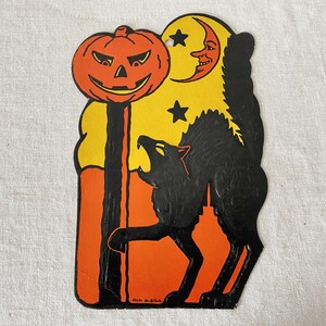 May include: A die-cut Halloween decoration featuring a black cat arching its back, a bright orange jack-o'-lantern on a post, and a yellow crescent moon with a face. The design includes two black stars against a yellow and orange background.