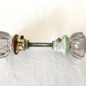 May include: Antique glass door knobs with a ribbed design. The knobs are clear with aged metal hardware and a connecting rod. The metal shows signs of wear with peeling paint and rust, giving them a vintage aesthetic.
