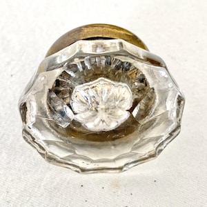 May include: Clear glass door knob with a faceted design and a brass base. The knob has a floral pattern in the center and is likely vintage. The glass is clear, allowing light to pass through.