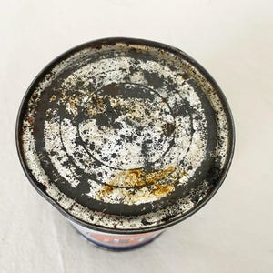 May include: A weathered metal can, showing signs of age and wear. The lid is marked with black, white, and rust-coloured patterns. The rim of the can is worn, indicating it is likely an antique or a vintage item.