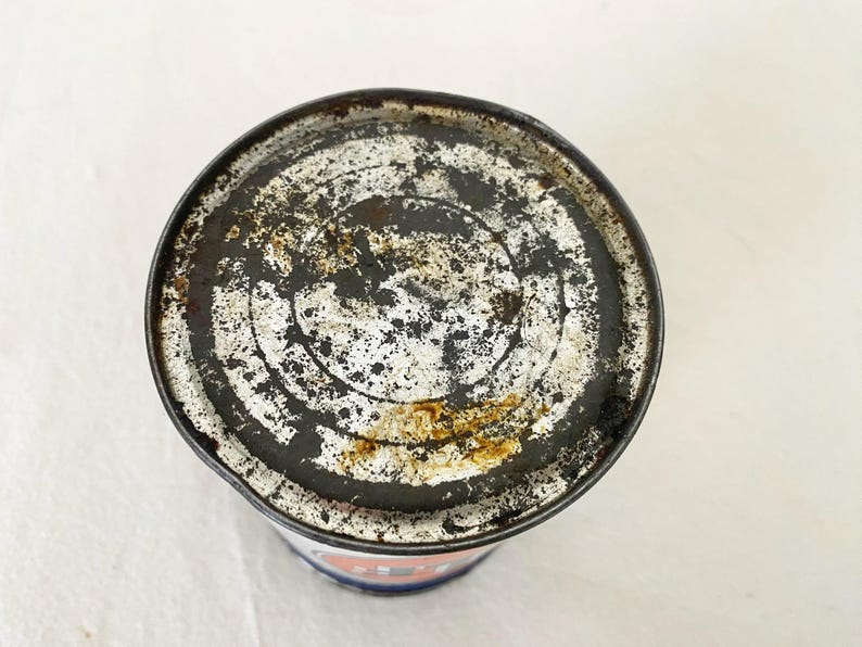 May include: A weathered metal can, showing signs of age and wear. The lid is marked with black, white, and rust-coloured patterns. The rim of the can is worn, indicating it is likely an antique or a vintage item.
