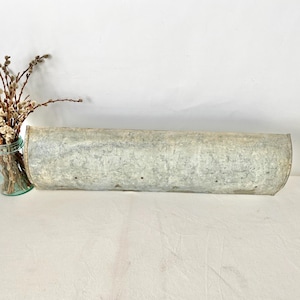 May include: A long, galvanized metal trough with a curved shape, likely for planting or decoration. The trough is silver-gray and has a weathered appearance. A small, turquoise glass jar with dried branches is to the left.