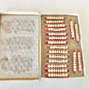 May include: An open cardboard box containing sets of artificial teeth. The teeth are arranged on orange and clear plastic strips. The left side of the box is filled with bubble wrap for protection. The teeth are a creamy white color.
