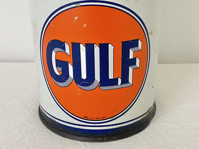 May include: A vintage metal can featuring the "GULF" logo in blue and white on an orange circle. The can has a white background with a blue and white border. The base of the can is black.