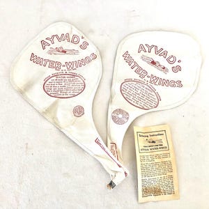 May include: A pair of vintage white canvas Ayvad's Water-Wings, a type of inflatable swim aid, with red lettering and a small swimmer illustration. The wings are deflated and folded. A small card with swimming instructions is included.