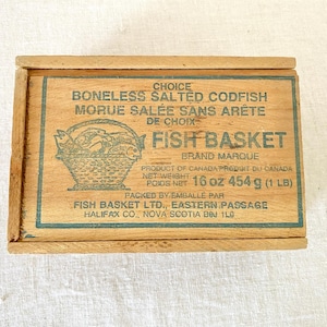 May include: Wooden box with a hinged lid. The box features text in English and French, including "FISH BASKET" and "BONELESS SALTED CODFISH." A green illustration of a basket with fish is in the center. The net weight is 16 oz (1 lb).