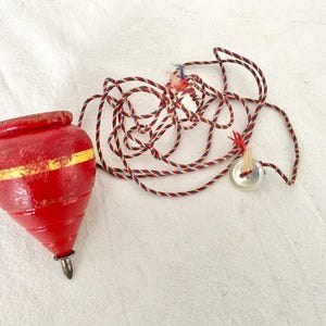 May include: A vintage red wooden spinning top with a yellow stripe, a metal tip, and a twisted red, white, and blue string. A small metal button is attached to the string. The toy is a classic children's game.