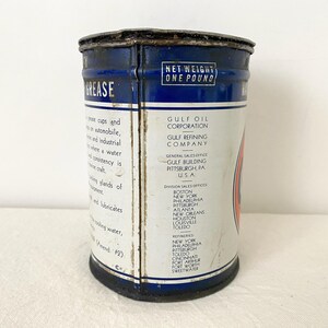 May include: A vintage Gulf Oil grease tin with a blue and white label. The tin is marked "NET WEIGHT ONE POUND" and lists the Gulf Oil Corporation's general sales office in Pittsburgh, PA, USA. The cylindrical tin shows signs of wear.