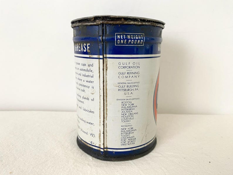 May include: A vintage Gulf Oil grease tin with a blue and white label. The tin is marked "NET WEIGHT ONE POUND" and lists the Gulf Oil Corporation's general sales office in Pittsburgh, PA, USA. The cylindrical tin shows signs of wear.
